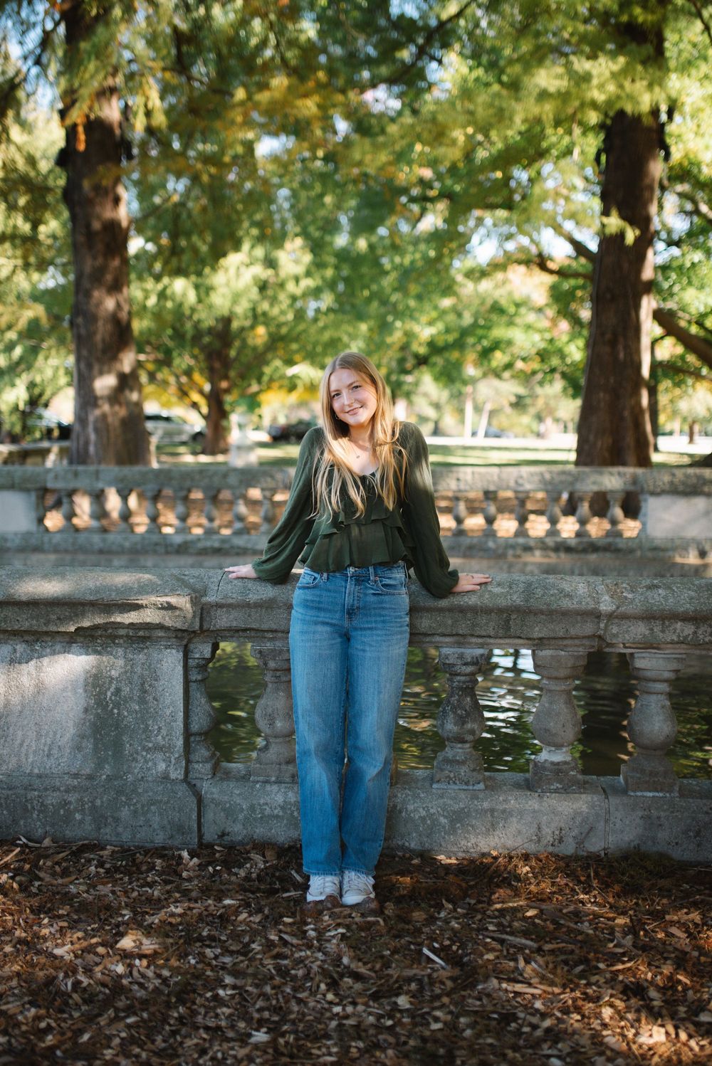 Person in blue jeans sits on stone wall in autumn park with fallen leaves and dappled sunlight.