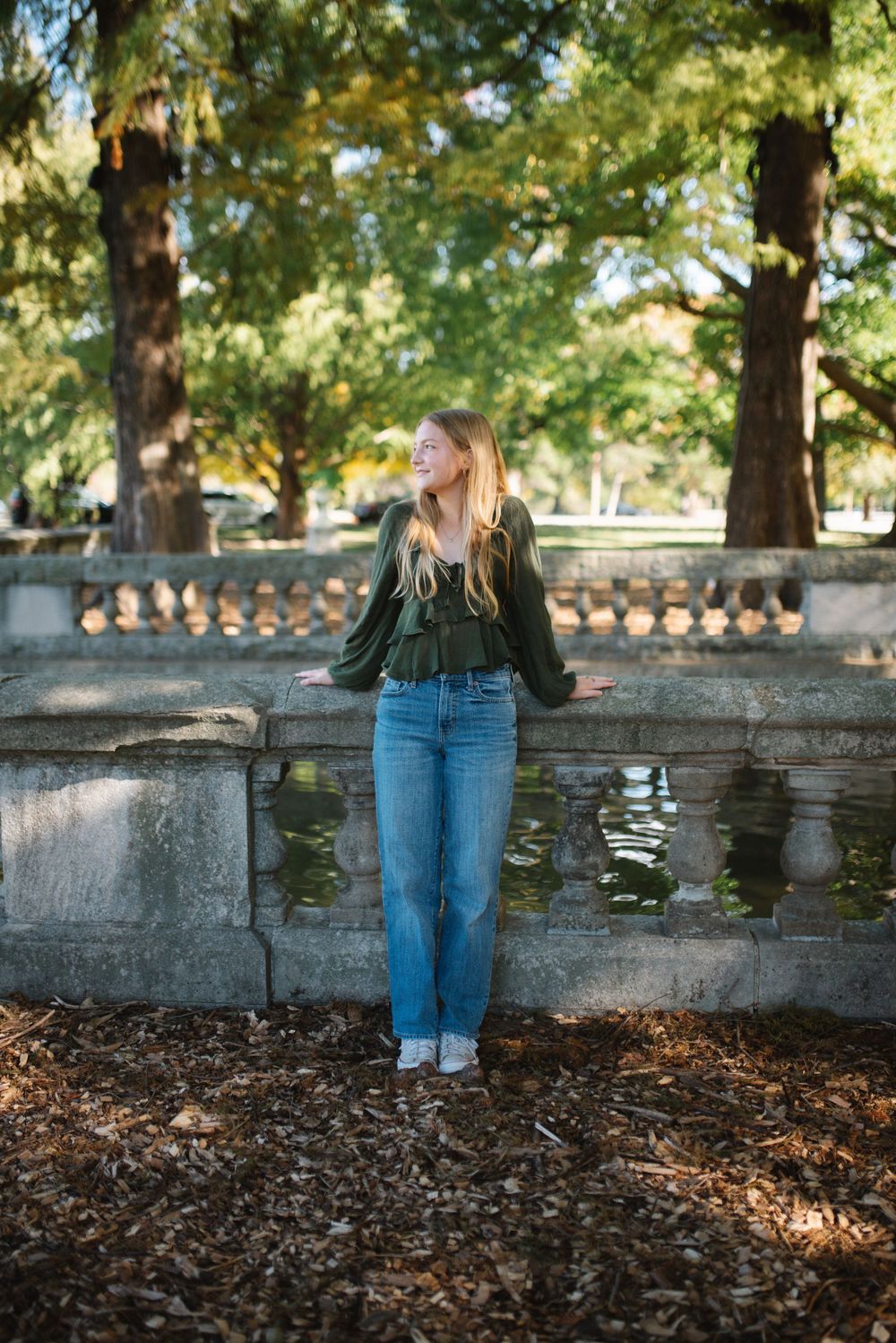 Person in casual fall outfit leaning against stone wall surrounded by autumn foliage in park setting.