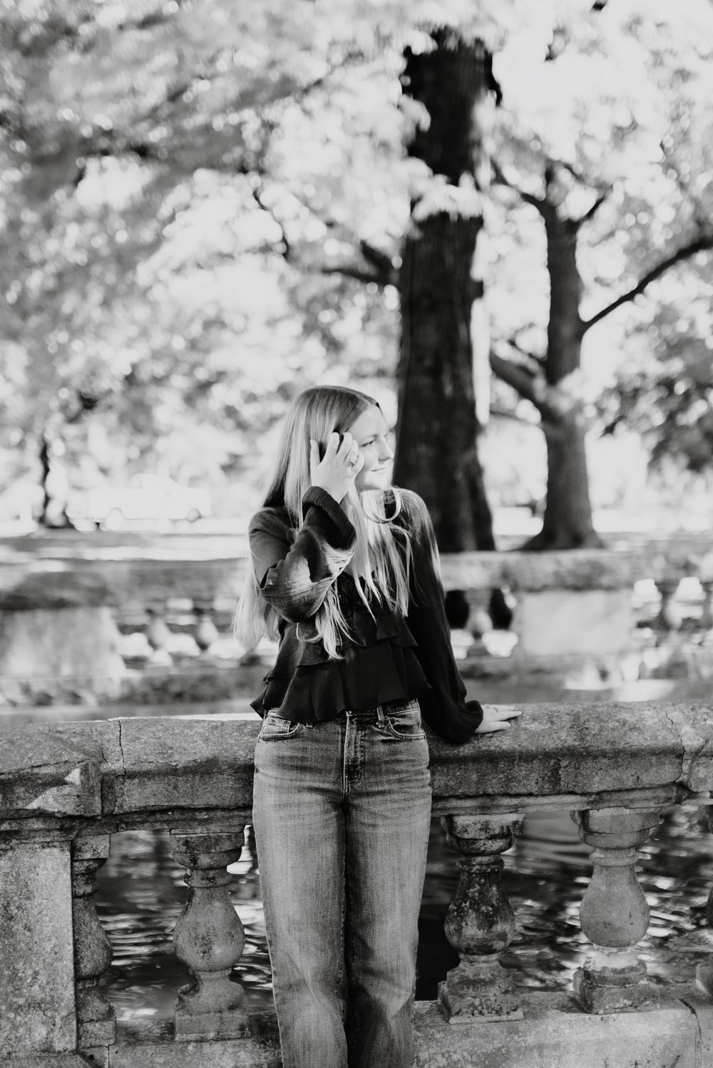 Black and white portrait of someone in casual attire leaning against stone wall in park setting.