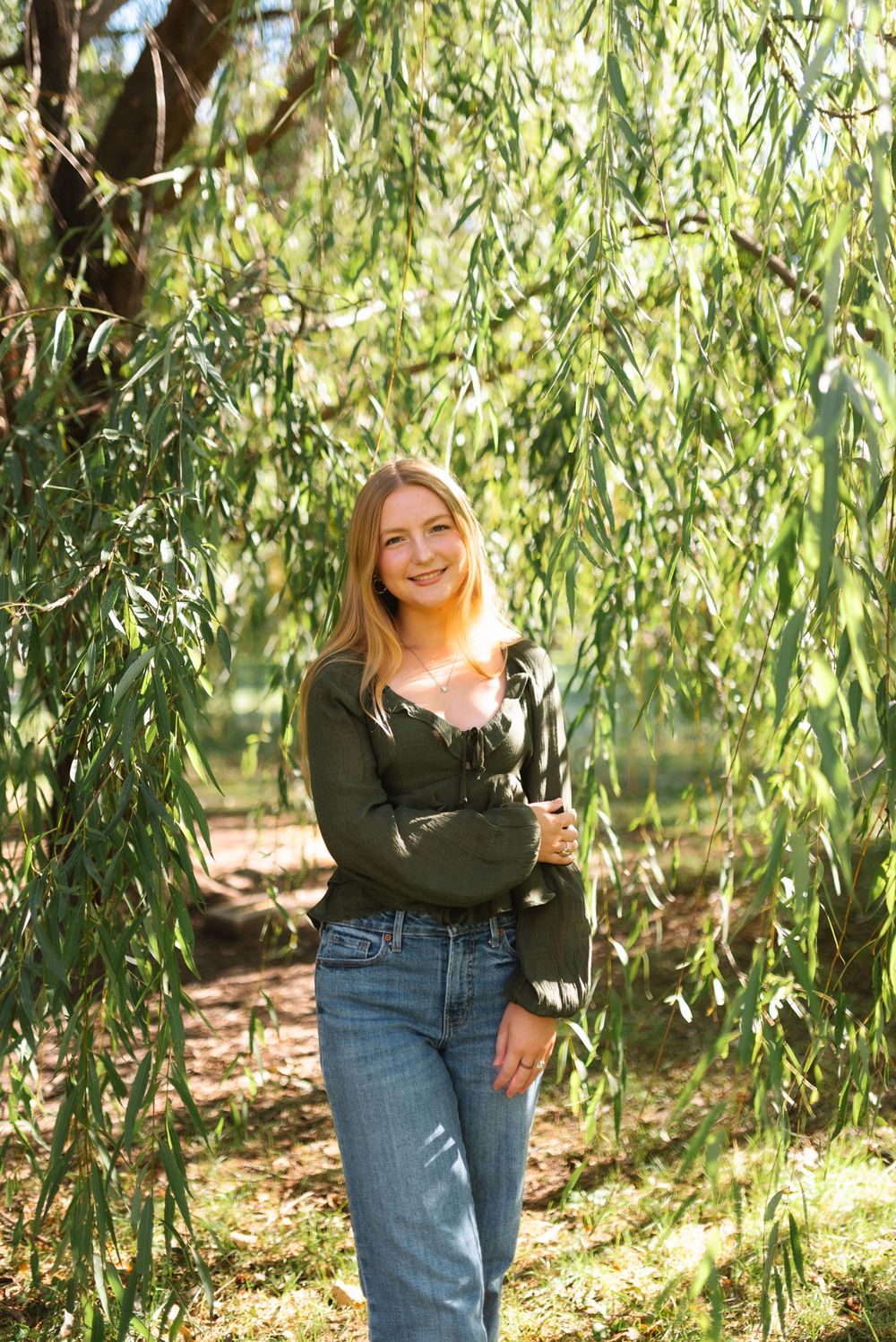 A person in a dark green sweater and jeans stands beneath cascading willow tree branches on a sunny day.