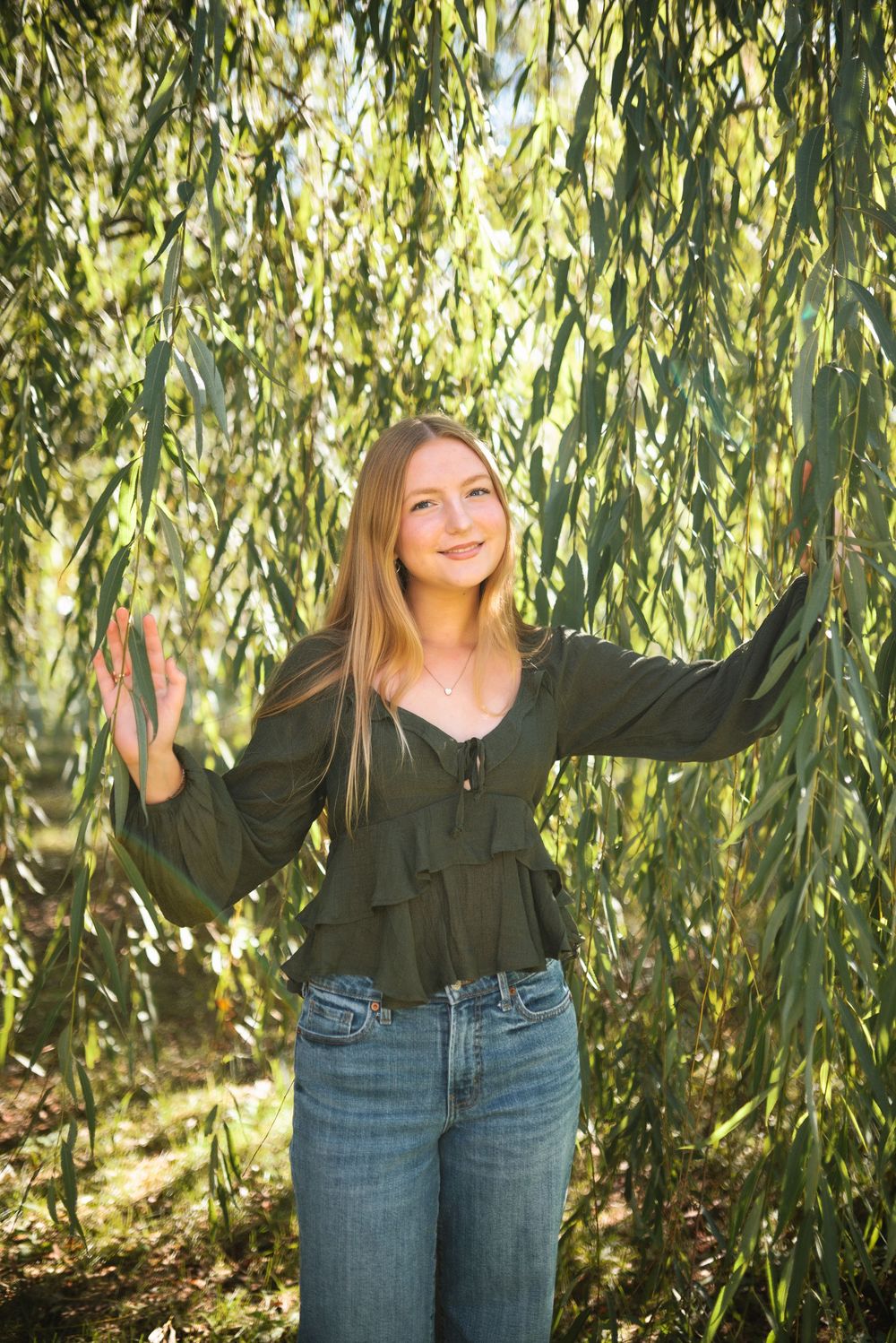 A person in a black ruffled top and jeans poses playfully under a weeping willow tree in a series of shots.