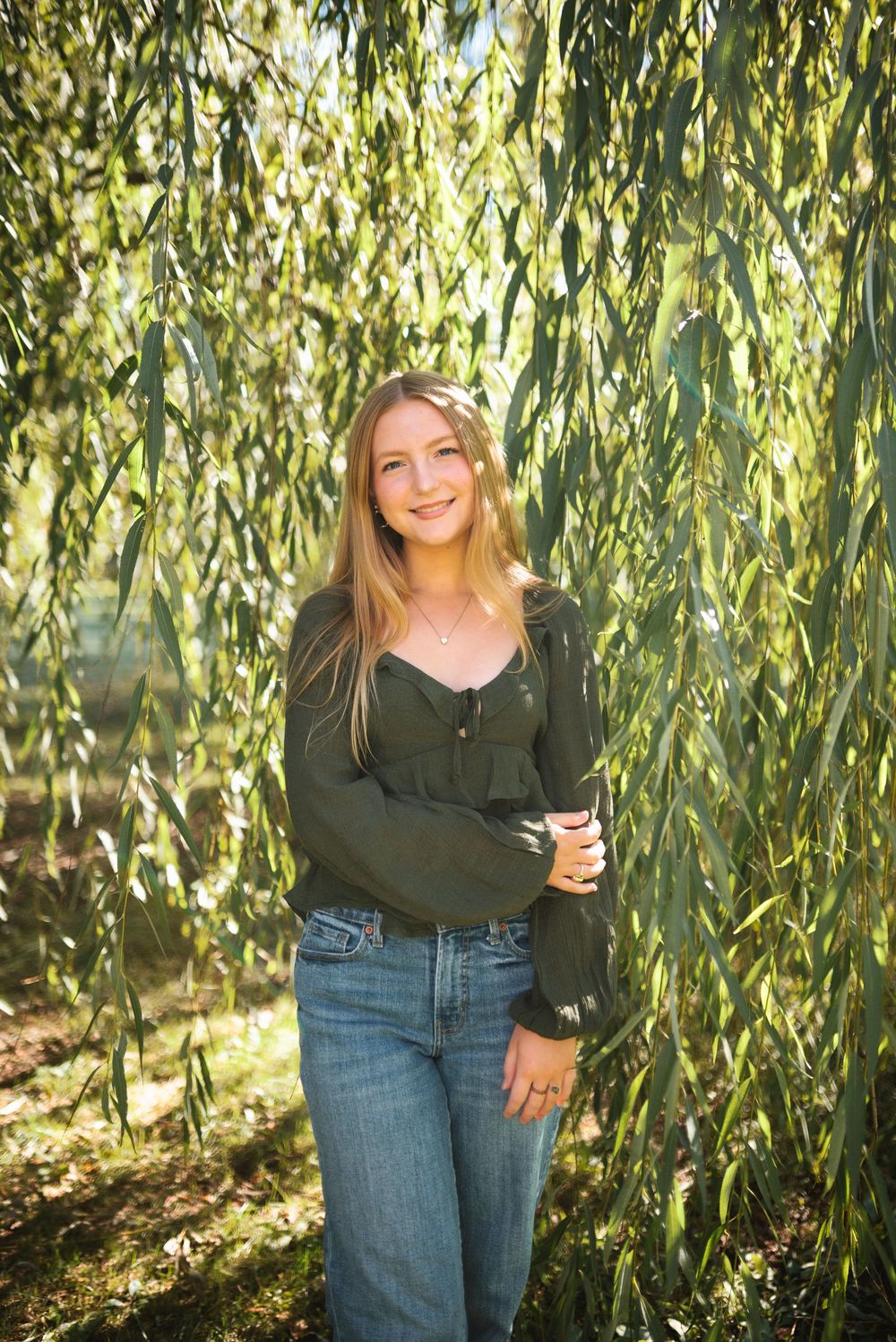 Portrait of someone in a green sweater standing under flowing willow tree branches in sunlit natural setting.