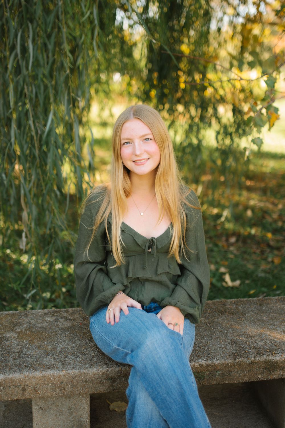A person in a green top and jeans sits on a stone bench under a willow tree on a sunny day.