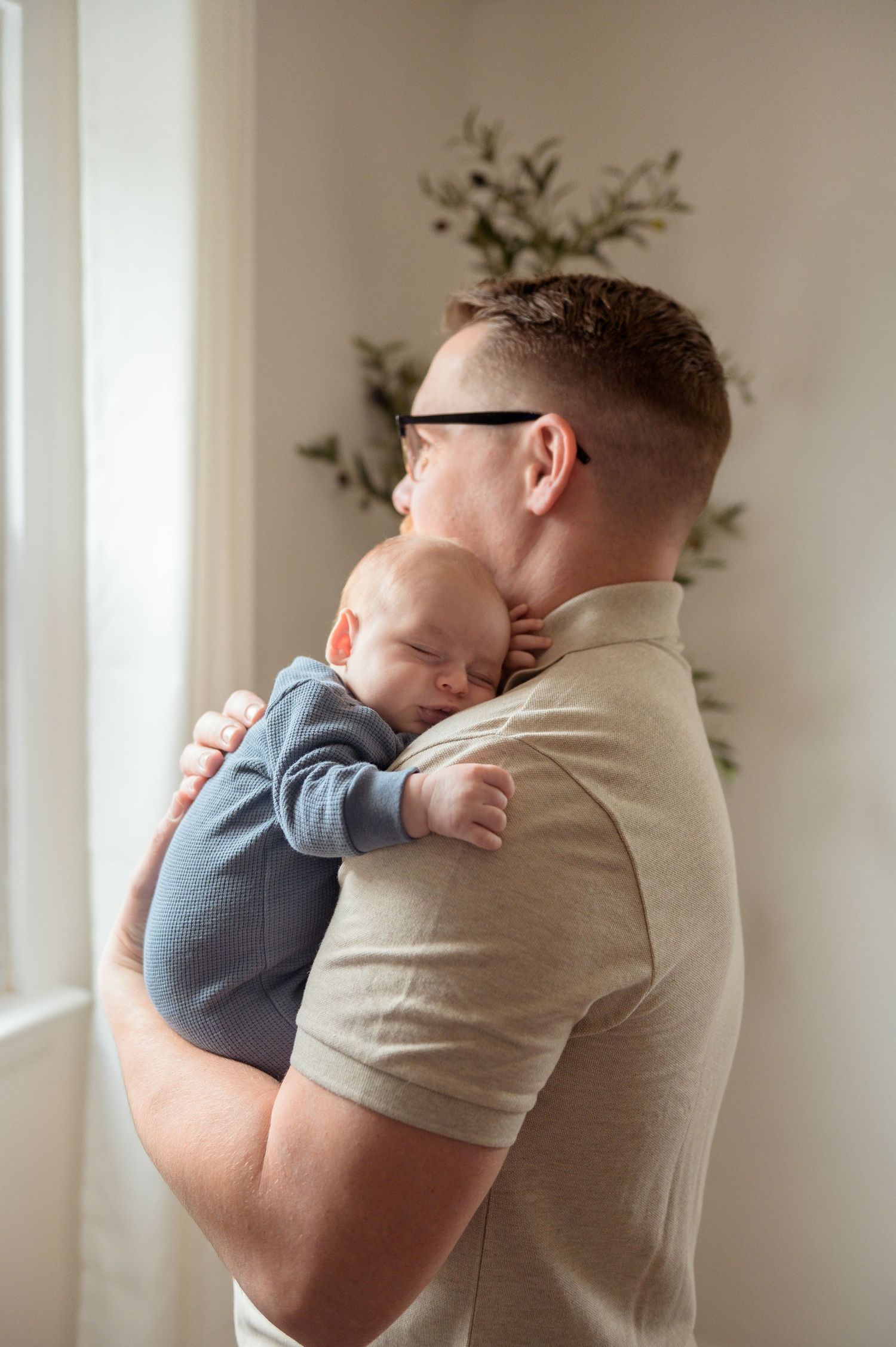 A tender moment captured as a baby in blue sleeps peacefully on their parent's shoulder near a window.