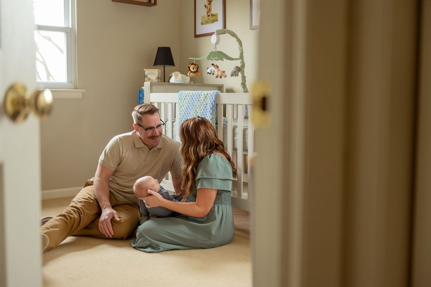 Two people sit on the floor of a nursery room holding and gazing lovingly at a newborn baby.