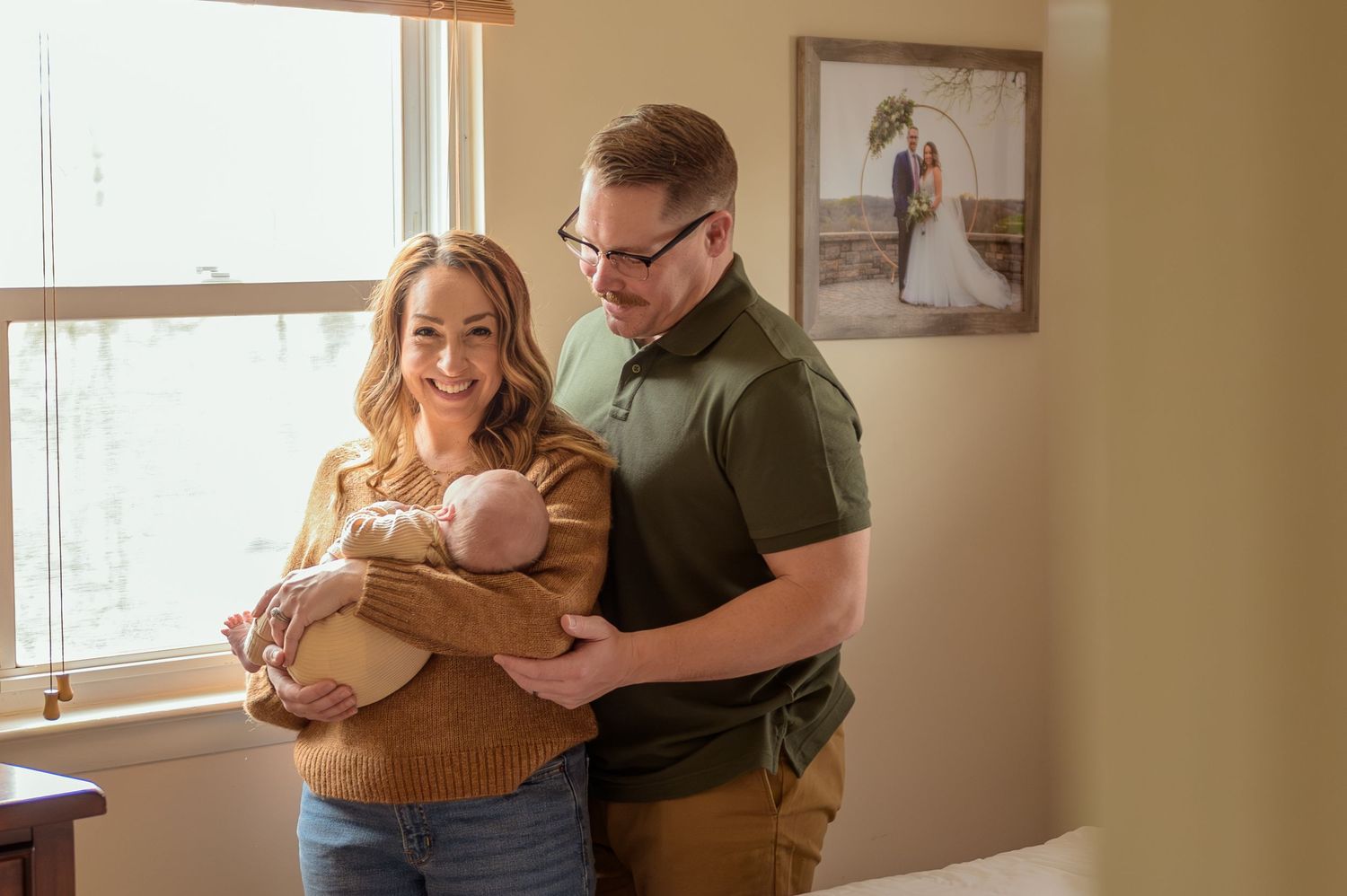A couple stands by a window in a cozy home interior holding their newborn baby while sharing an intimate moment together.