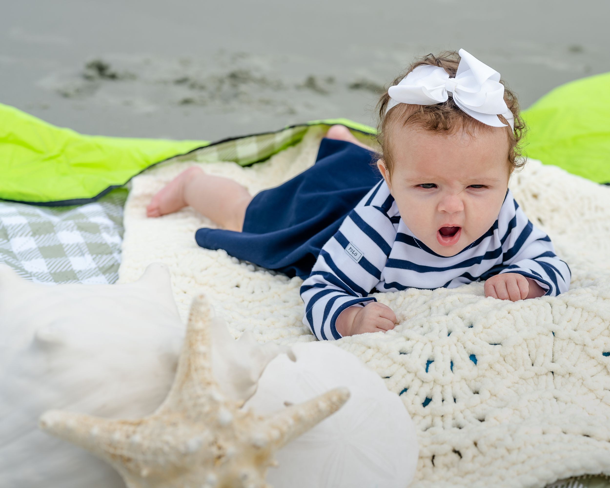 A baby in a striped shirt and white hat plays in a sandbox with a plastic shovel and bucket.