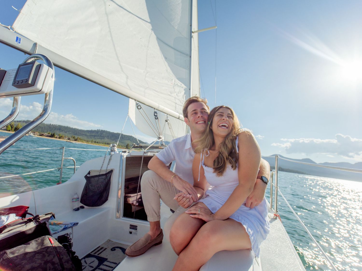 Happy couple sitting together on a sailboat deck, enjoying a sunny day on turquoise waters with white sails above.