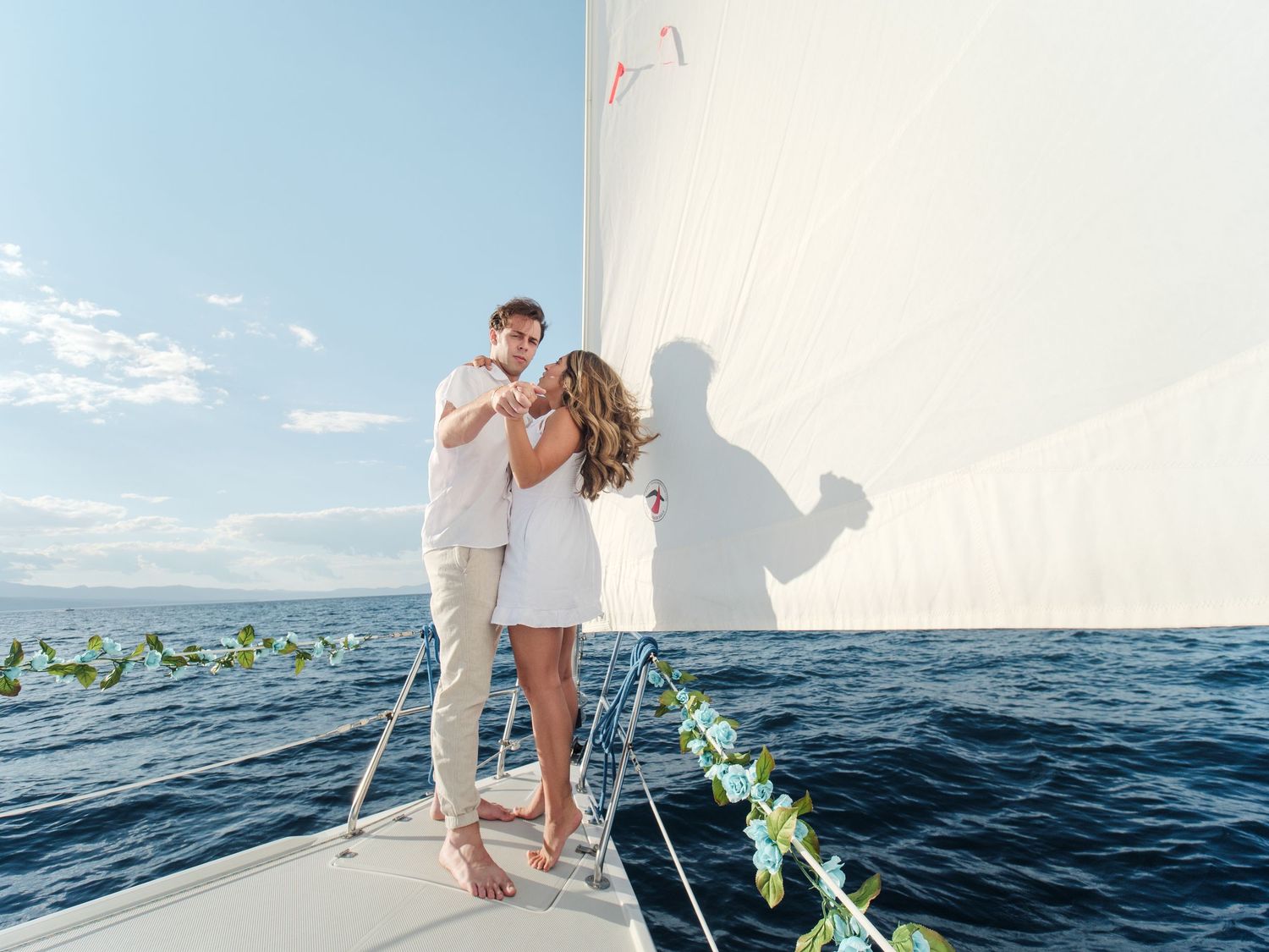 Romantic couple embracing on a decorated sailboat deck with white sails and blue ocean backdrop.