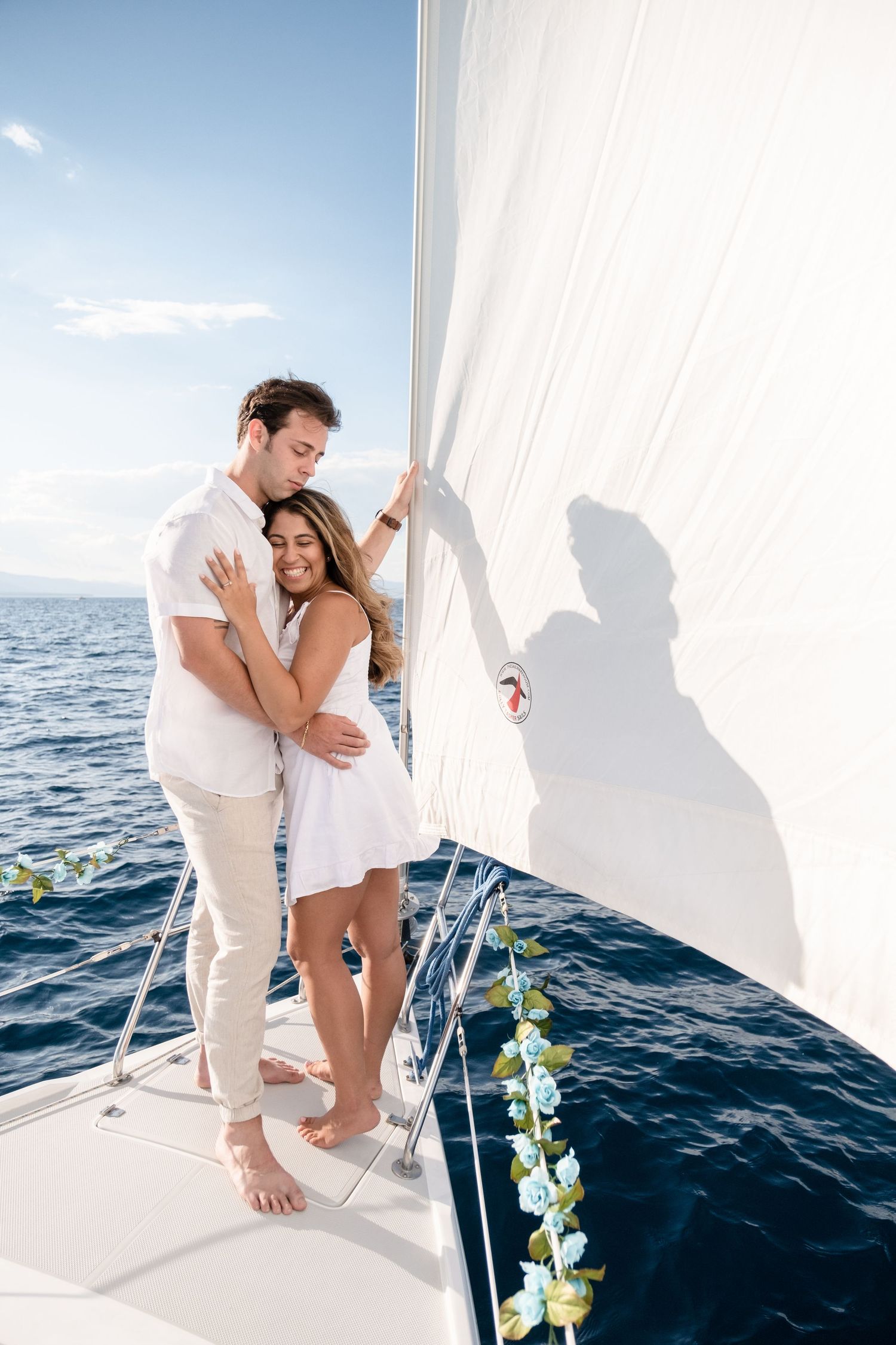 A romantic couple embraces on a sailboat deck, dressed in white, with the blue ocean in the background.