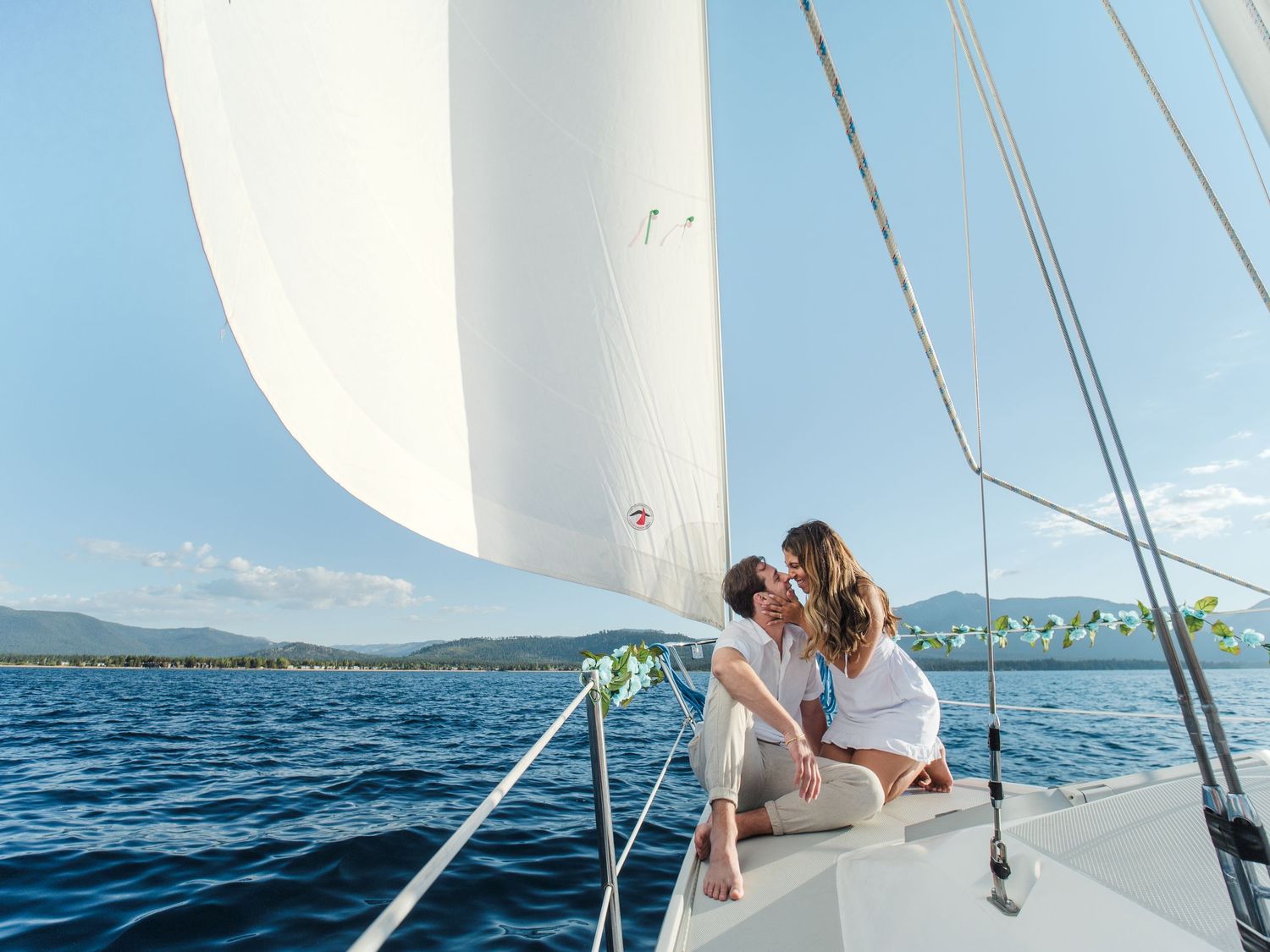 Romantic couple kissing on the bow of a white sailboat on calm blue waters with green hills in the background.