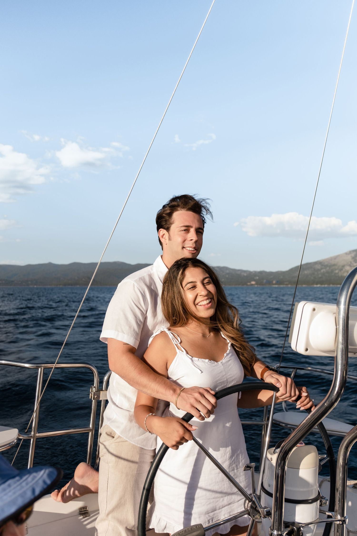 A smiling couple steers a sailboat together on calm blue waters with scenic mountains in the background.
