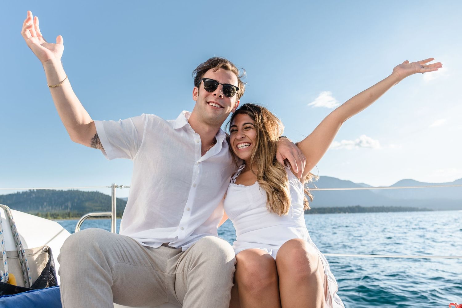 Happy couple with arms raised sitting on a boat deck with blue ocean and mountains in the background on a sunny day.