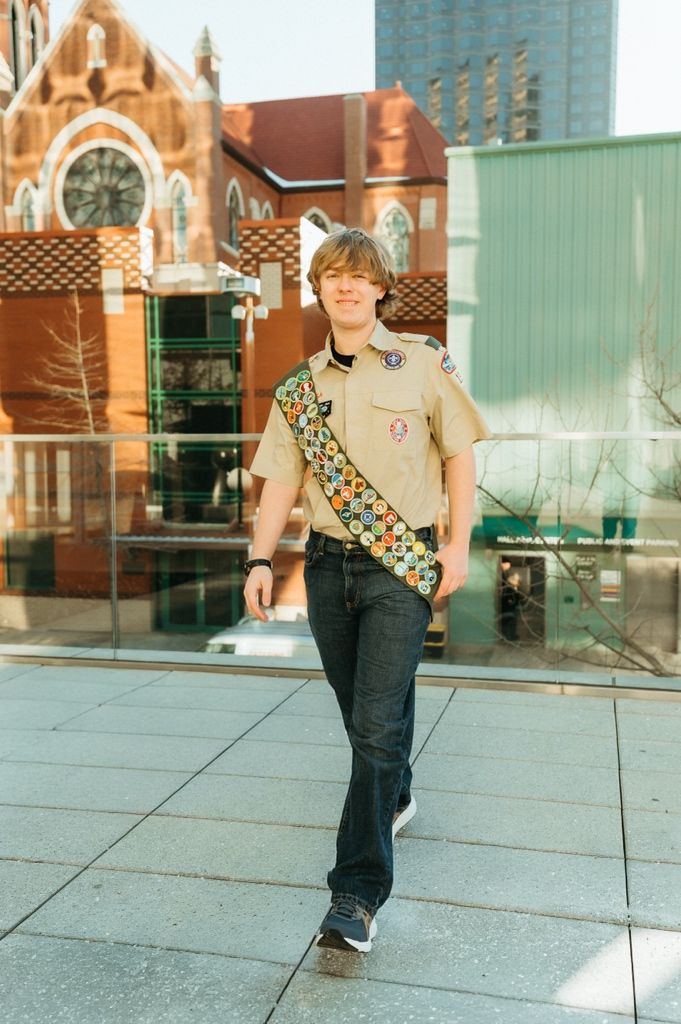 Scout in uniform walks along glass-walled building exterior in urban setting.