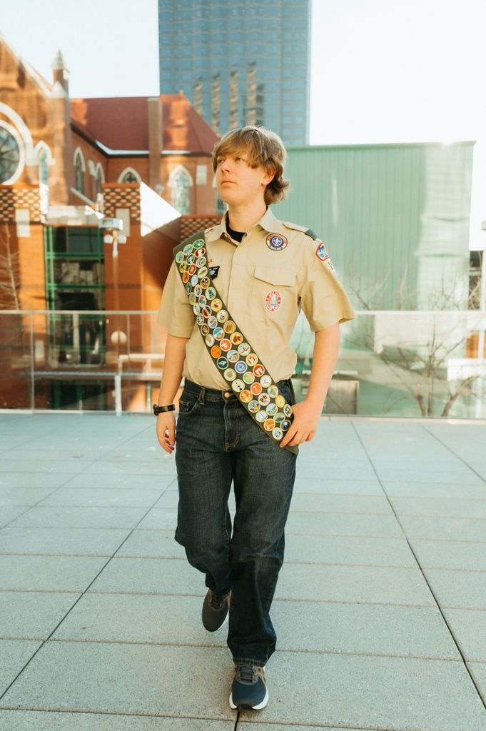 A Scout in uniform wearing a sash full of merit badges stands on an urban rooftop plaza.