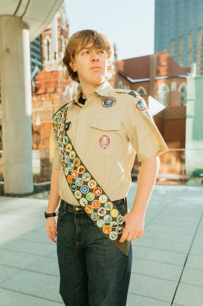 Scout displaying merit badge sash and uniform stands against modern building backdrop.