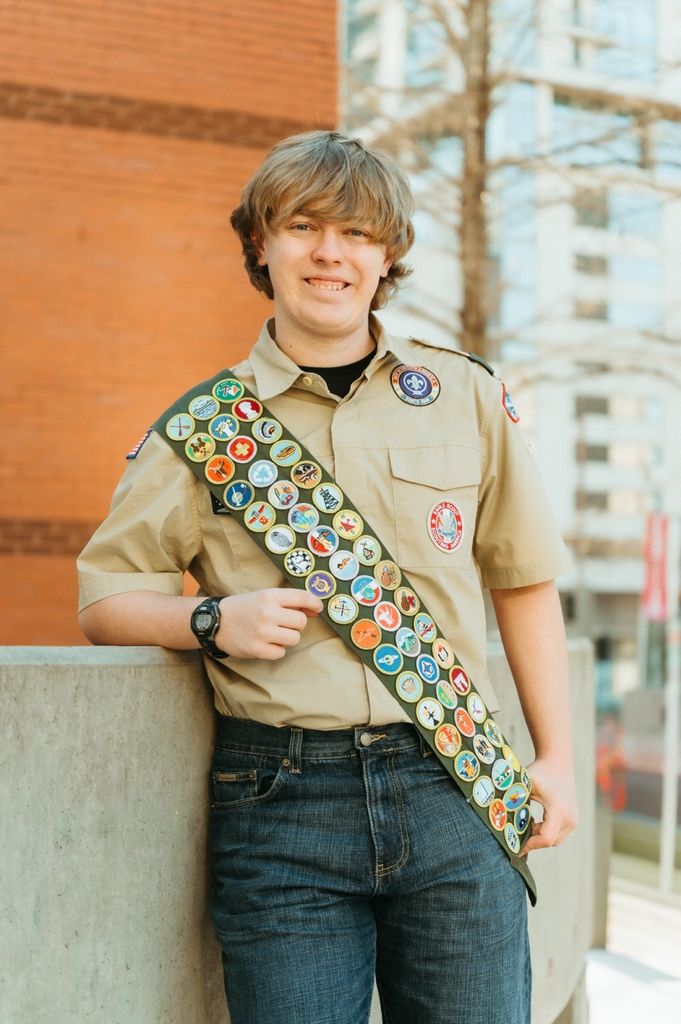 Scout wearing tan uniform shirt and jeans displays colorful merit badge sash with multiple badges against brick wall.