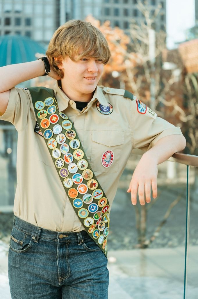 A scout uniform with merit badges and patches displayed on the tan shirt and sash against a blurred background.