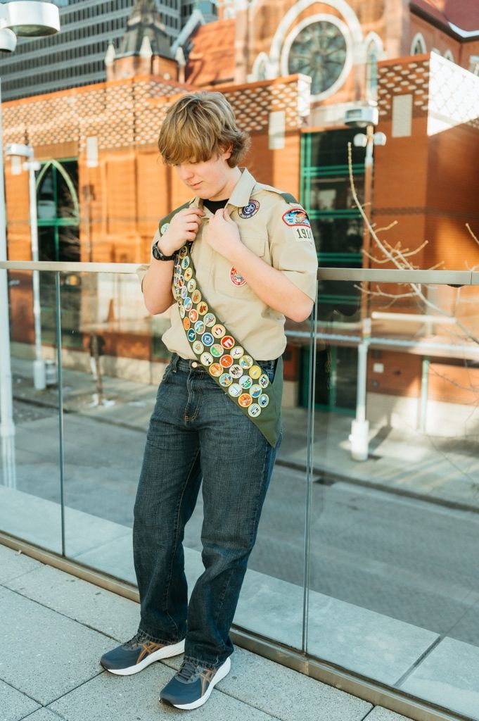 Scout in uniform with merit badge sash poses against brick building and glass railing.