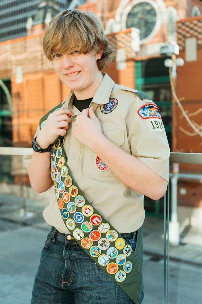 A scout wearing a khaki uniform shirt displays colorful merit badges on their sash.