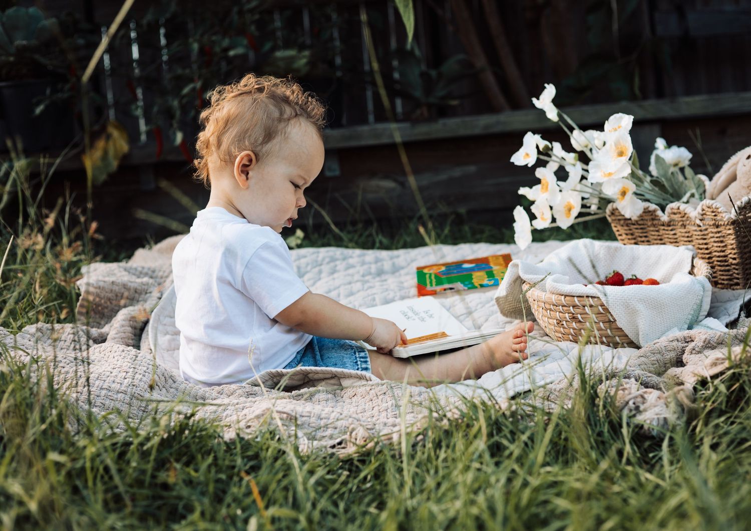 A toddler sits on a blanket in a garden, reading a book at a charming outdoor picnic with flowers and fruit.