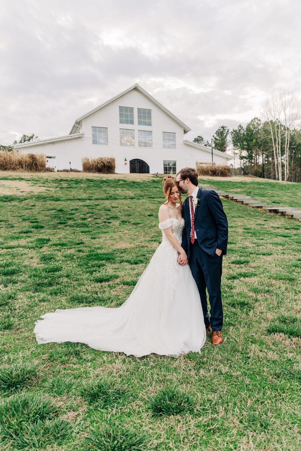 Couple in wedding attire walking together on grassy hill with white farmhouse buildings in background.