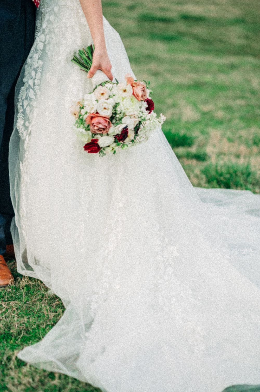 Flowing white lace wedding dress train spread out on green grass with delicate floral bouquet.
