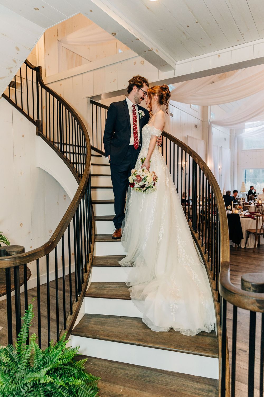 A couple in wedding attire shares a romantic moment on a curved wooden staircase with black railings and white walls.