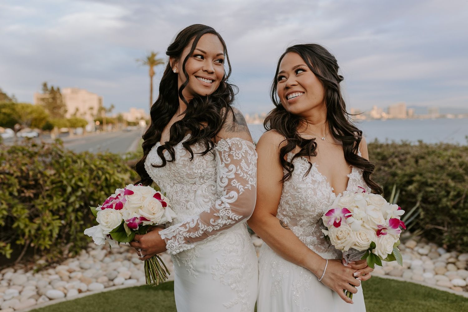 Two brides in white wedding dresses share a joyful moment together while holding floral bouquets outdoors with the San Diego cityscape.