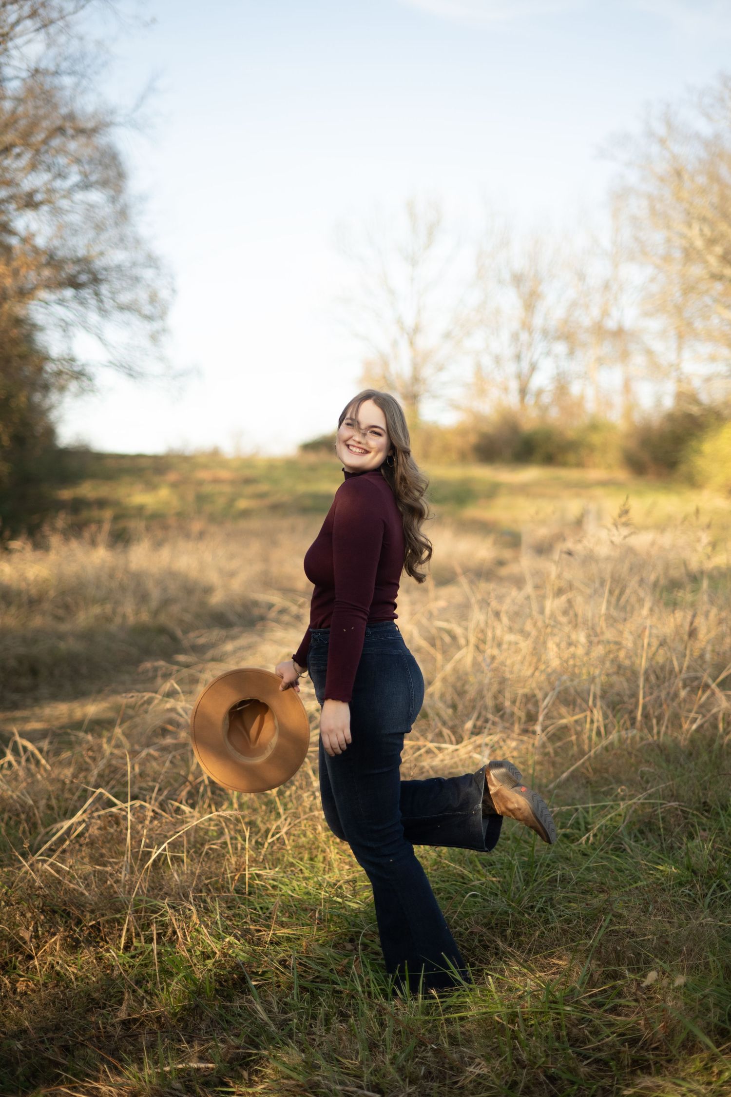 A figure stands in a sunlit field at golden hour wearing a burgundy sweater and holding a brown hat while striking a playful pose.