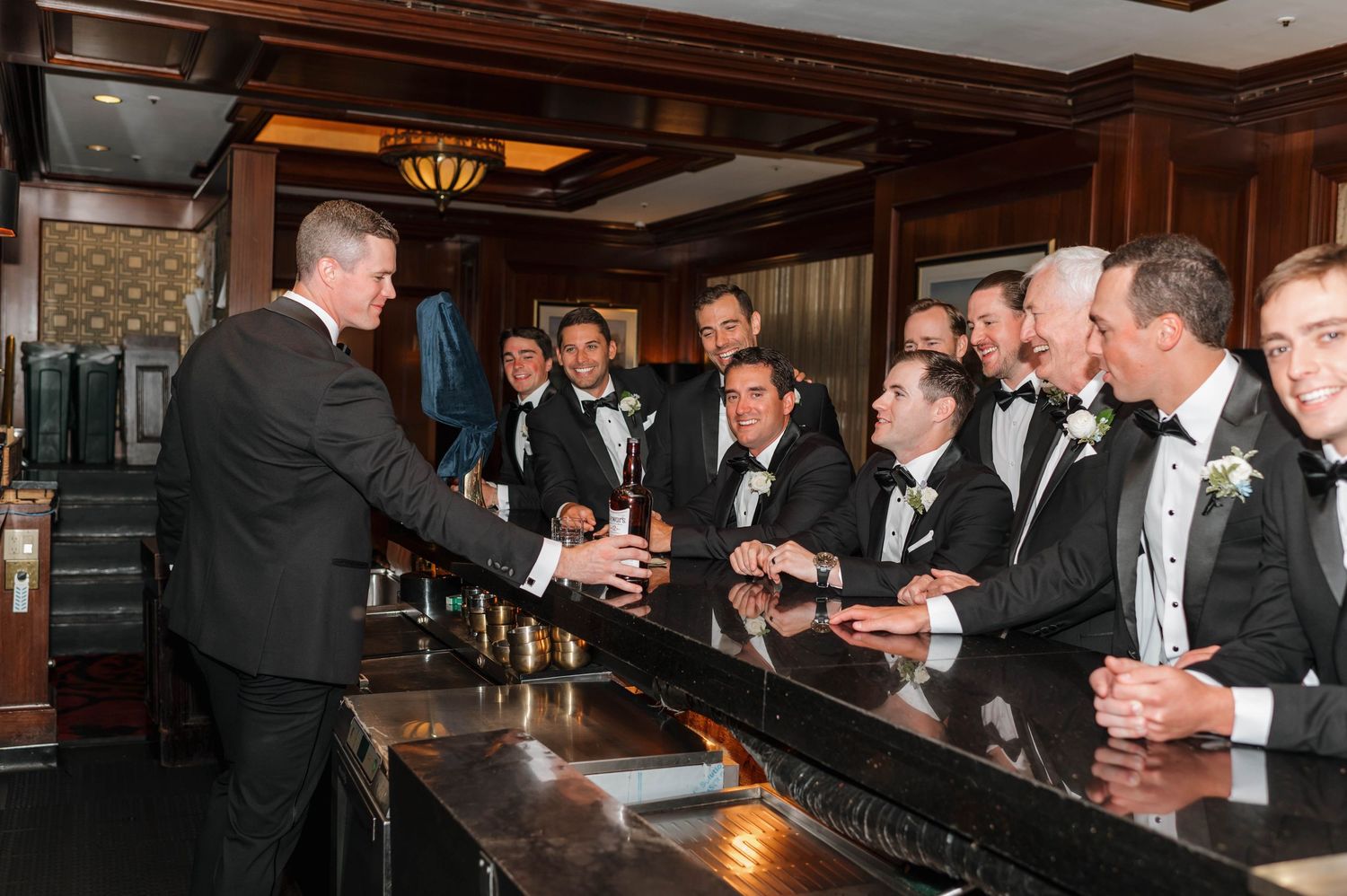 A group of men in formal attire gather around a dark wooden bar counter during what appears to be a social event.
