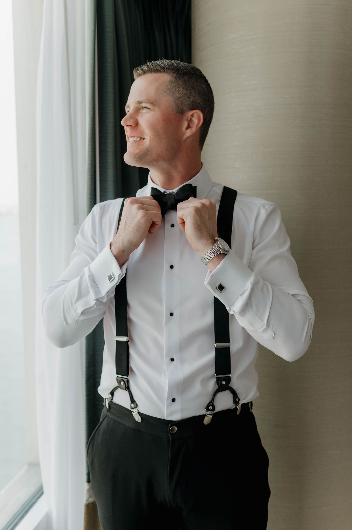 A person in a white dress shirt and black suspenders adjusts their collar while looking to the side near a window.