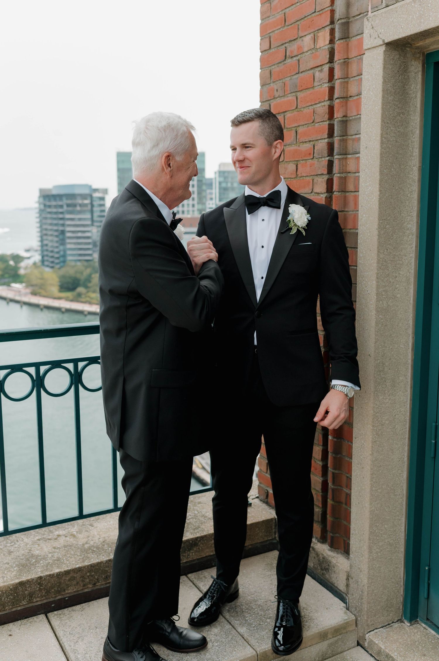 Two men in black tuxedos share a tender moment on a balcony overlooking a city skyline.