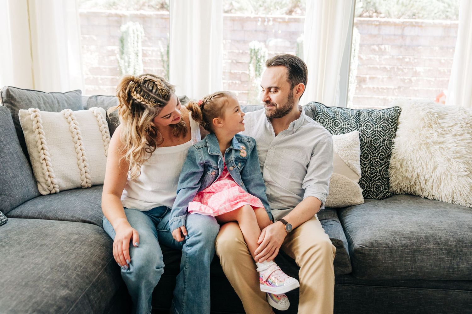 A Charlotte, NC family sits together on a gray couch in a bright room with exposed brick walls, sharing a tender moment.