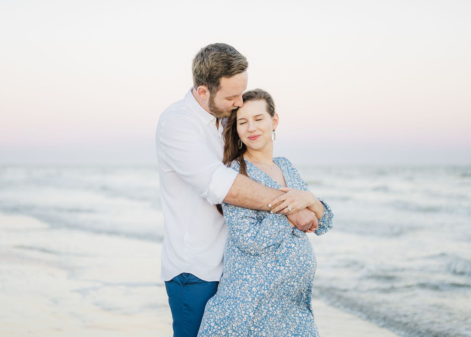 A romantic couple embraces on the beach at sunset with ocean waves and soft pink sky in the background.