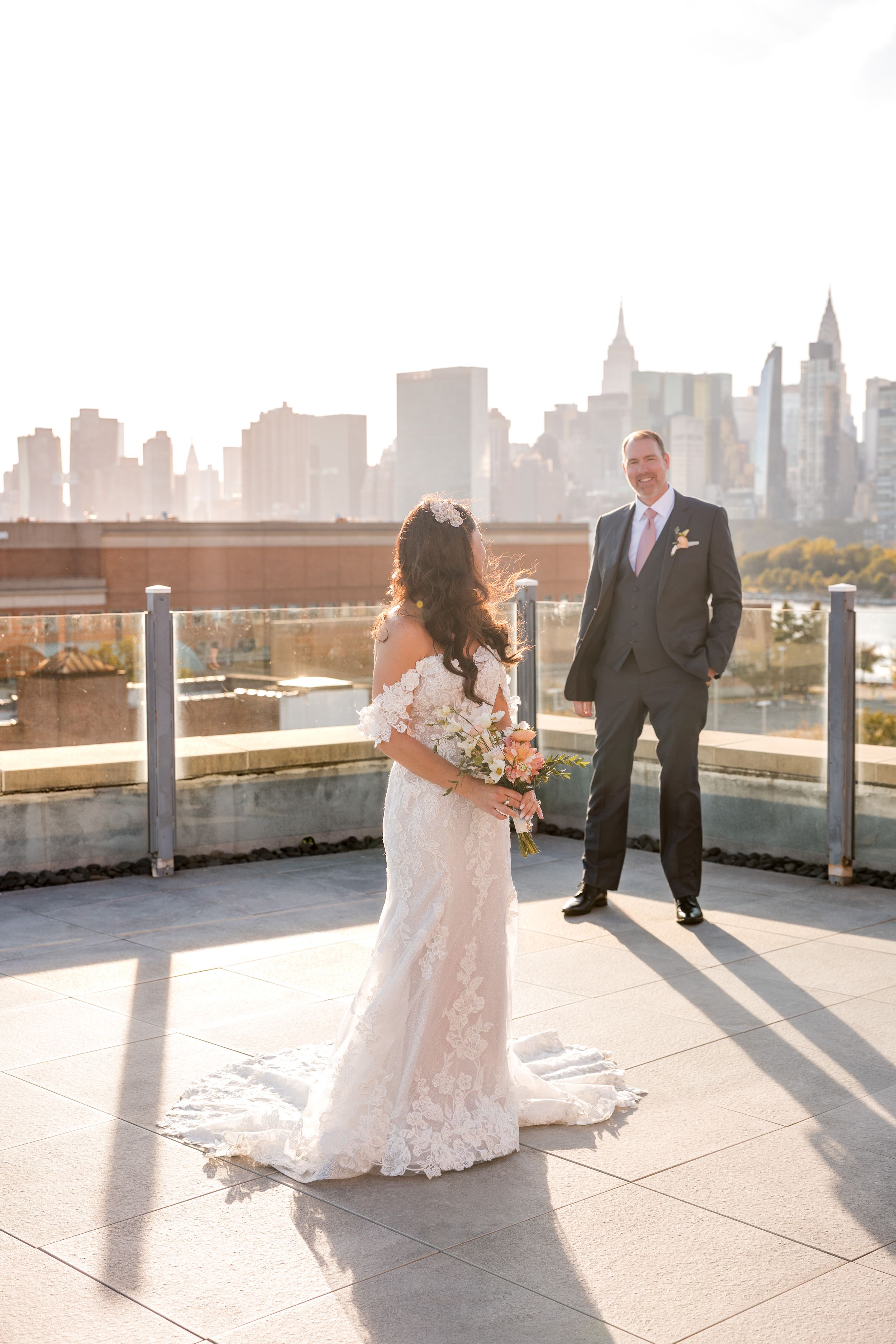 Bride and groom share a romantic first look on a rooftop terrace with the New York City skyline glowing behind them.