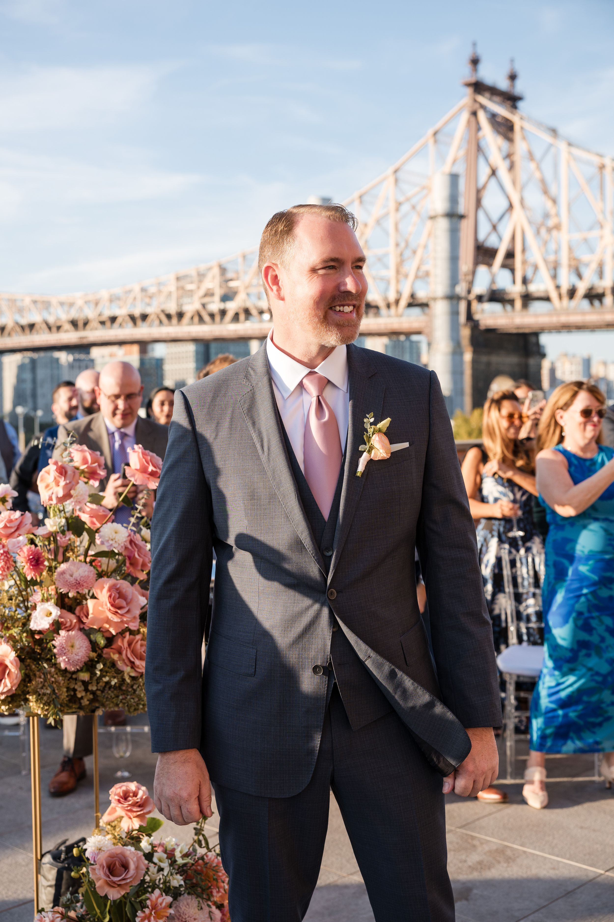 Groom in gray suit with pink tie smiling at outdoor wedding ceremony near Queensboro Bridge in New York City.
