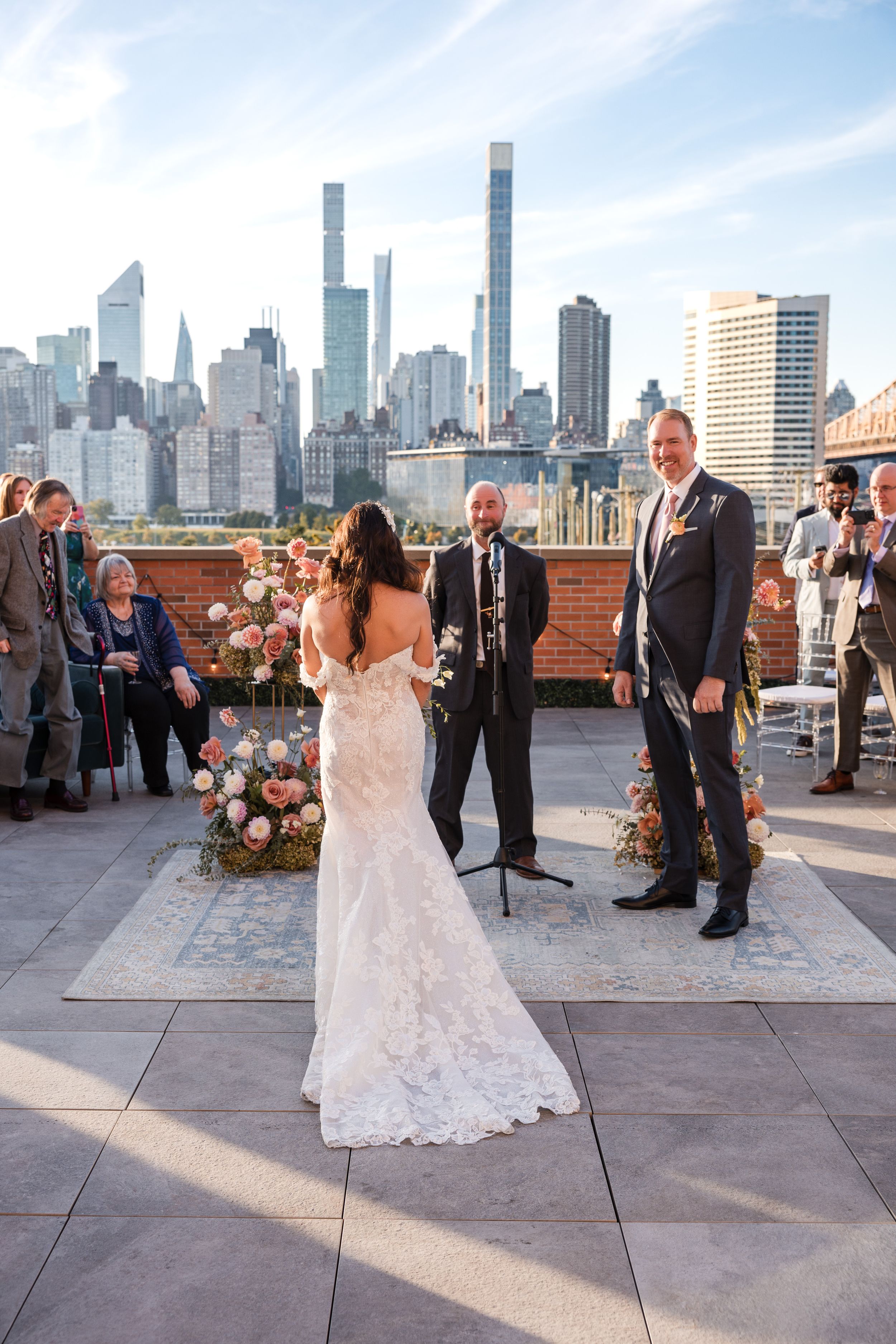 Rooftop wedding ceremony with NYC skyline backdrop, bride in lace gown facing groom in suit before officiant.