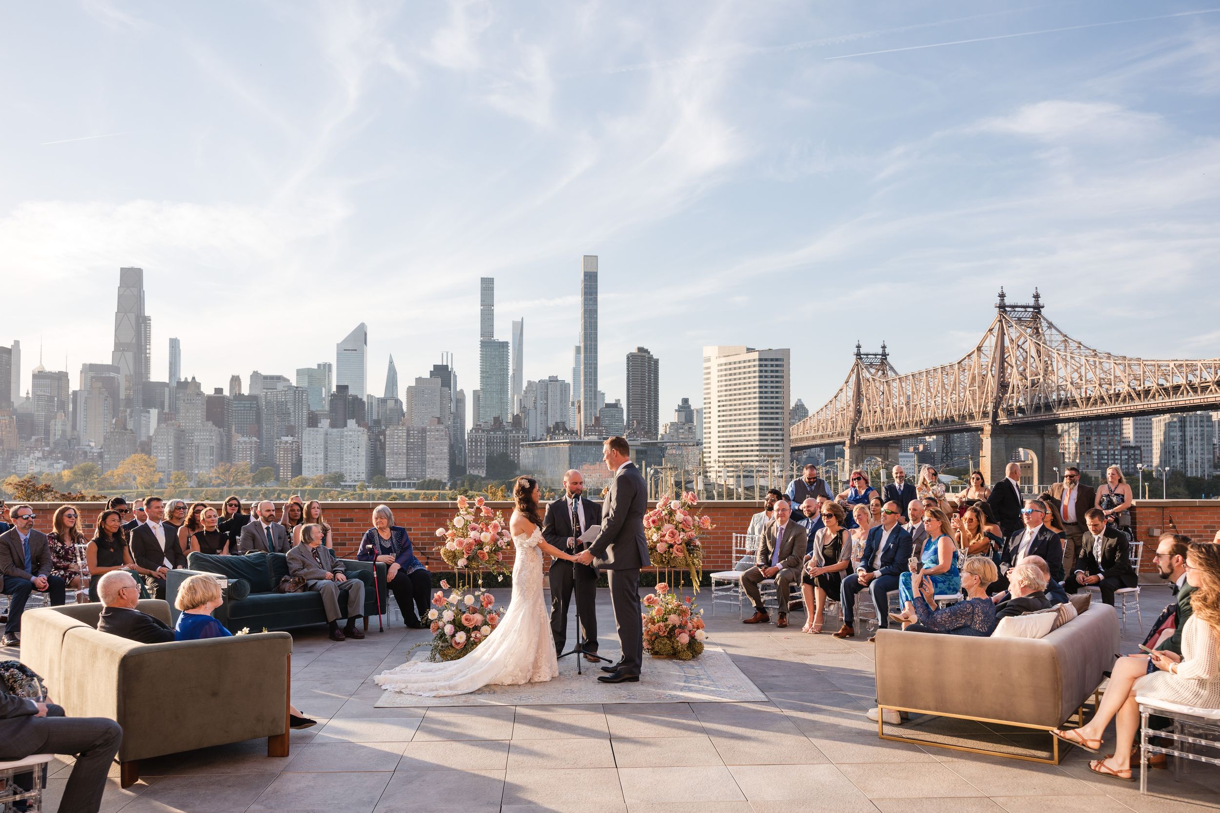 Rooftop wedding ceremony with NYC skyline and Queensboro Bridge backdrop, guests seated on stylish furniture under clear blue sky.