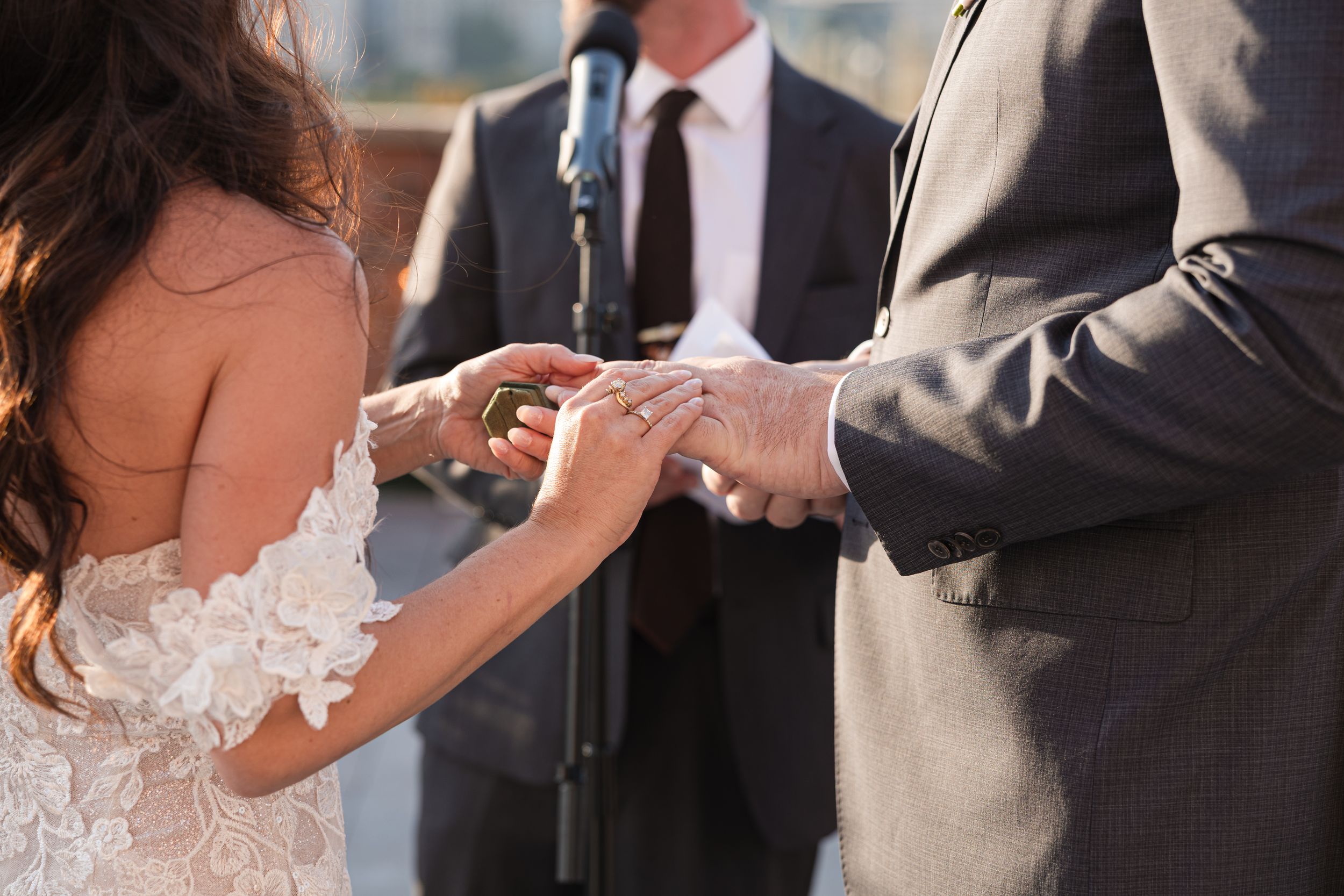 Bride placing wedding ring on groom's finger during outdoor ceremony, wearing lace dress with officiant in background.