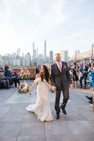 Bride and groom walk hand-in-hand after rooftop wedding ceremony with NYC skyline and Queensboro Bridge backdrop.
