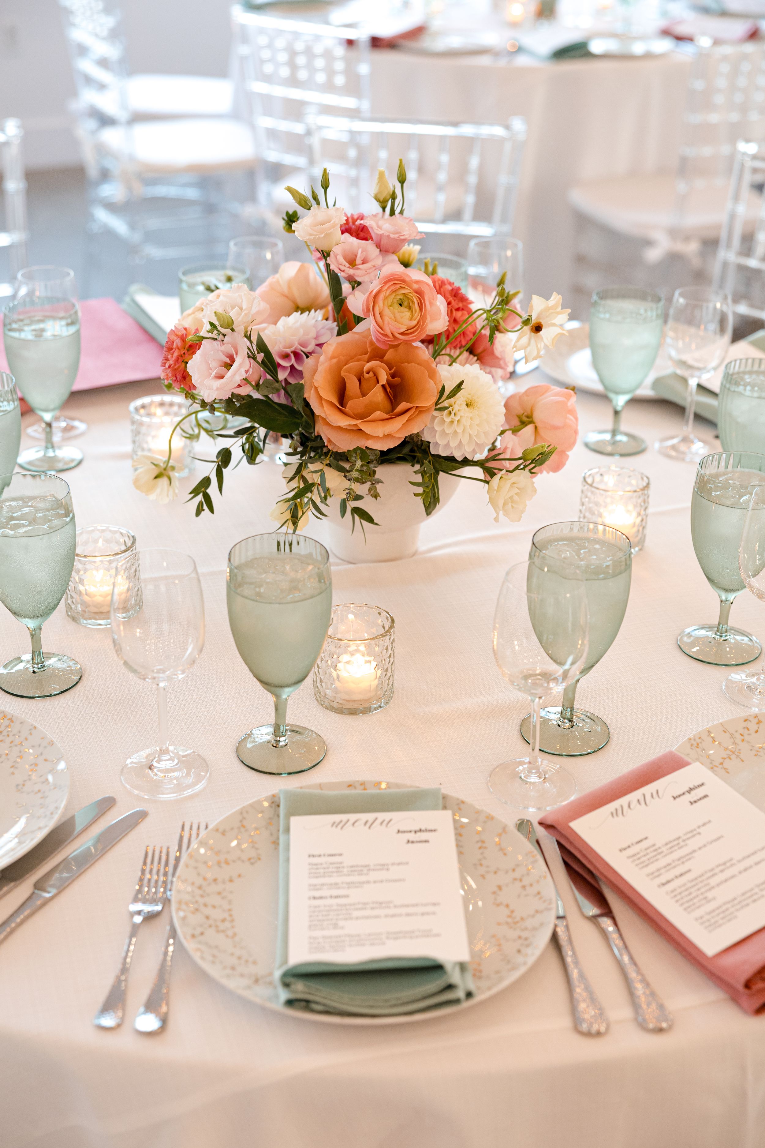 Elegant wedding reception table with coral floral centerpiece, mint green goblets, candles, and pink menu cards on white linen.