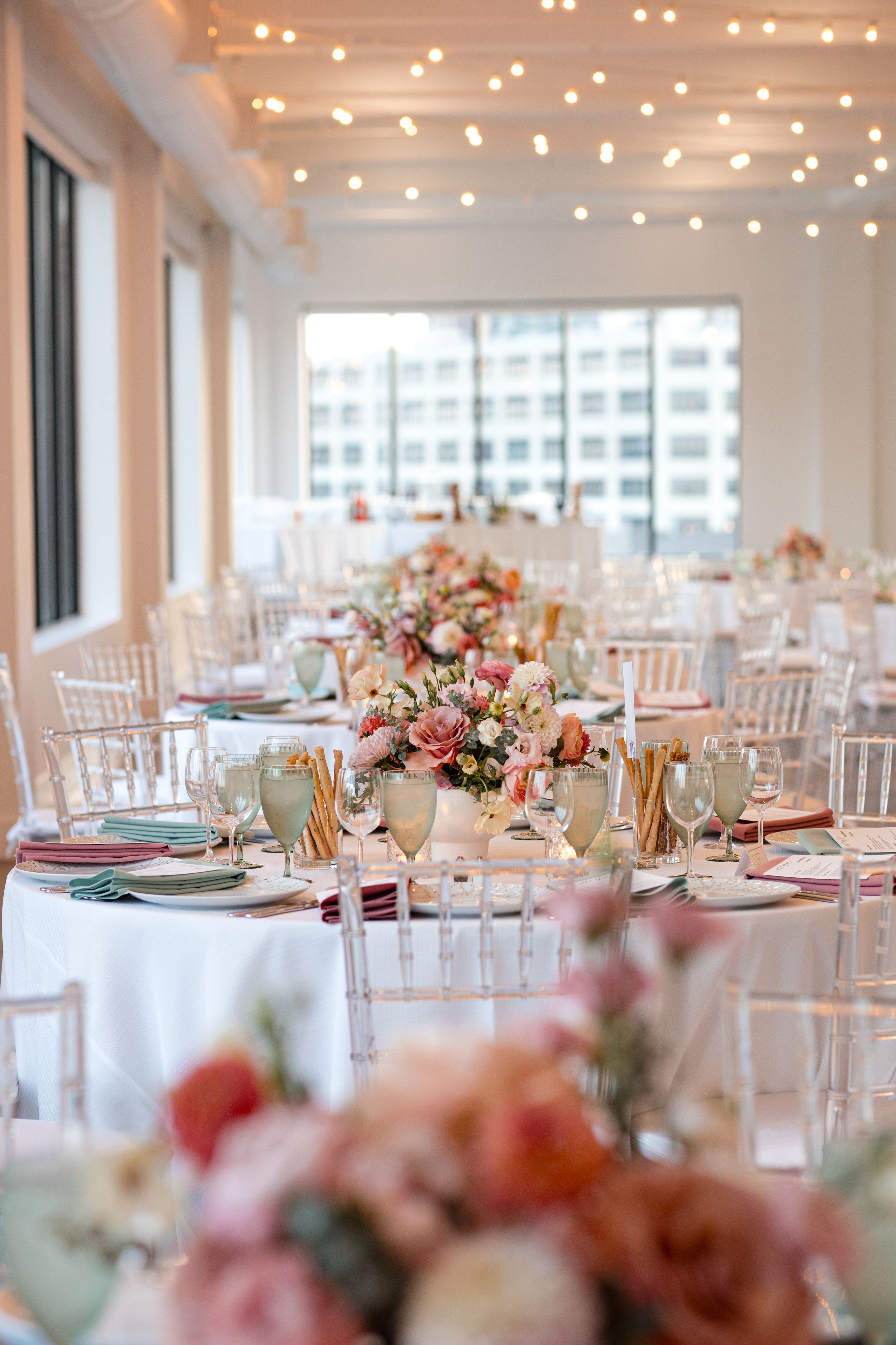 Elegant wedding reception with white tablecloths, pink floral centerpieces, chiavari chairs, and twinkling string lights overhead.