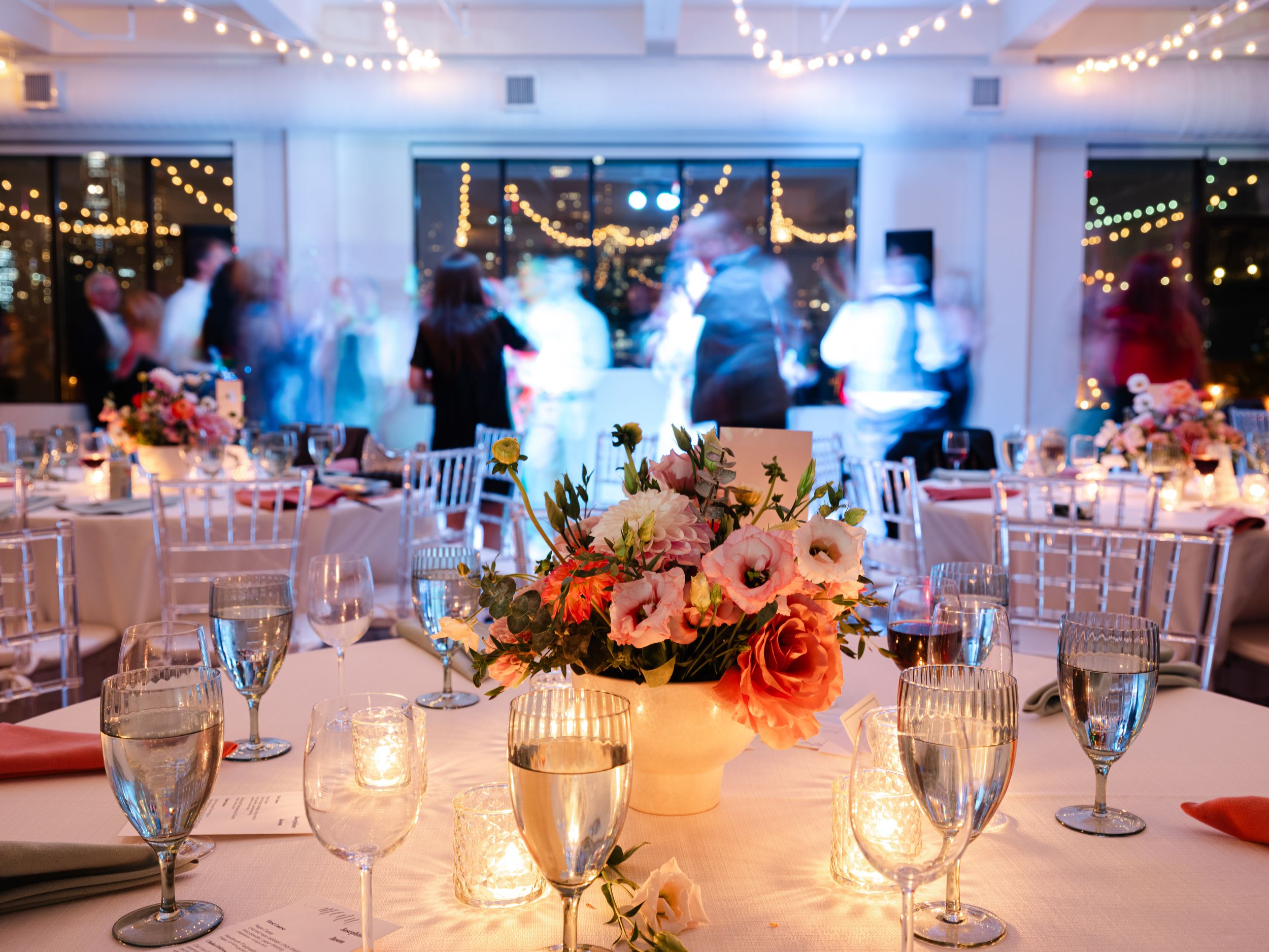 Elegant wedding reception with floral centerpiece, candlelit tables, glassware, and guests dancing under string lights.