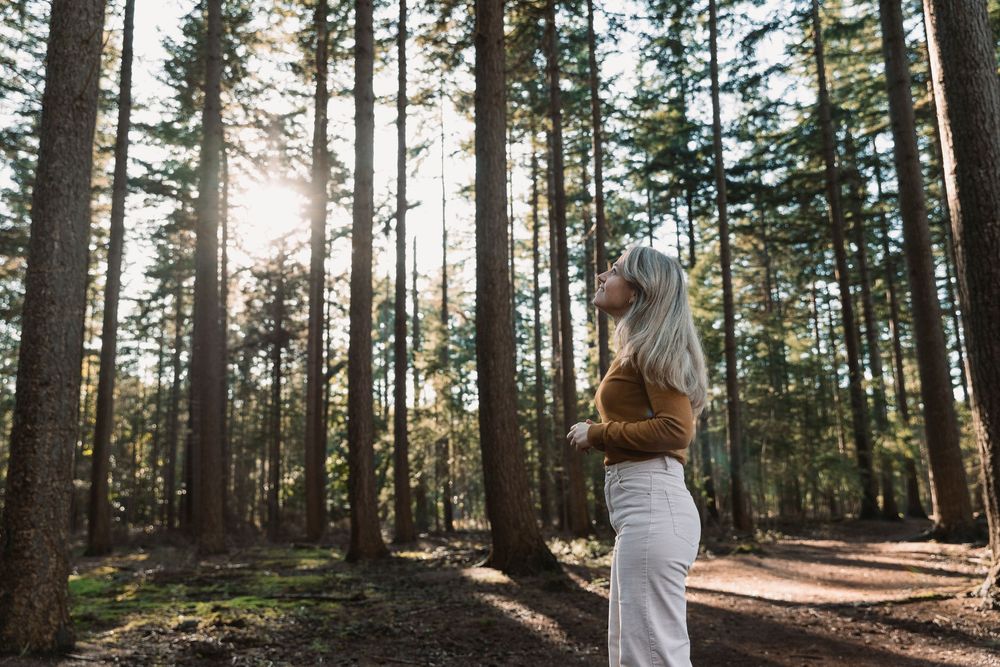 Person in brown sweater and white pants poses playfully in sunlit forest setting.