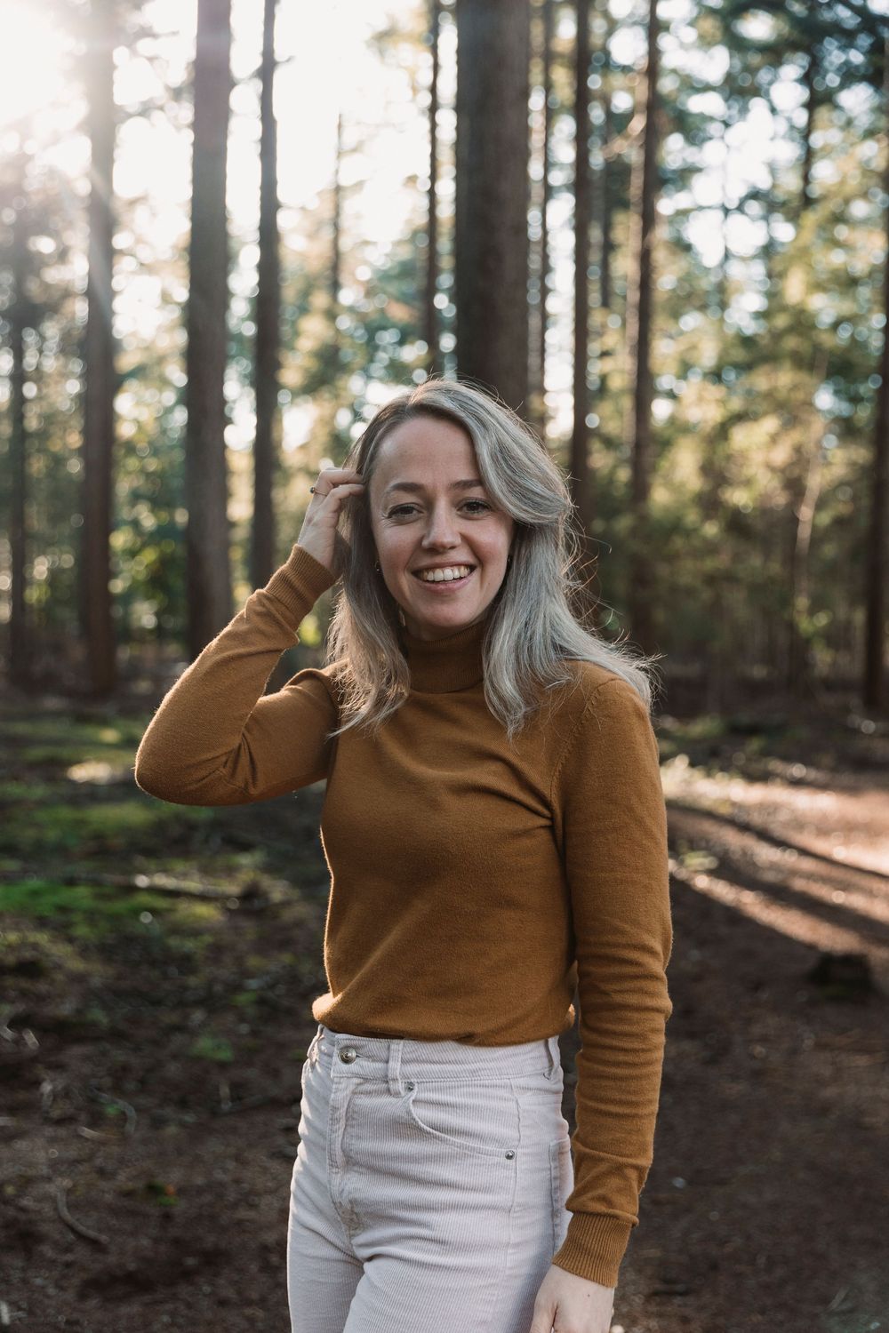 Figure wearing brown top and white pants stands among tall pine trees in dappled sunlight.