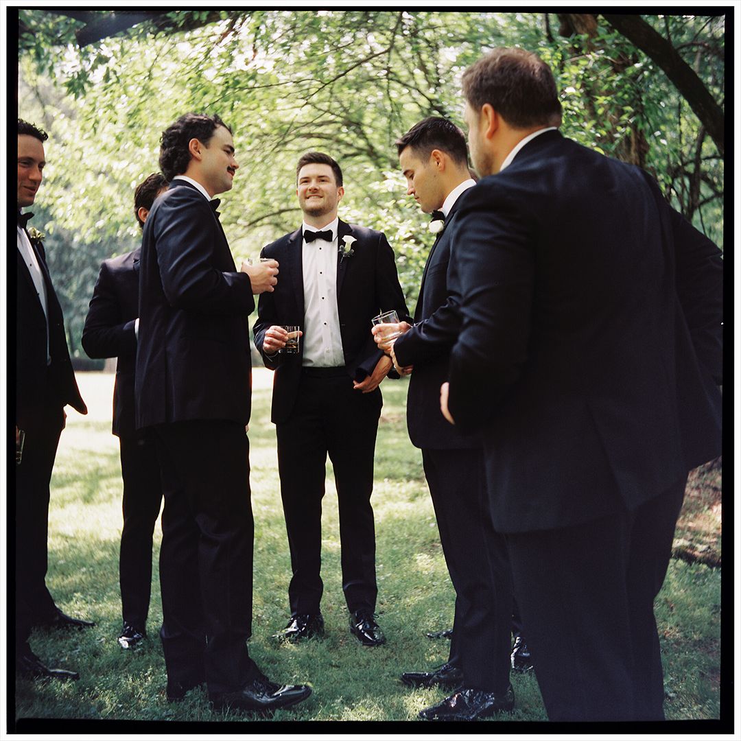 Groomsmen in black tuxedos share a lighthearted moment outdoors during a wedding photoshoot.