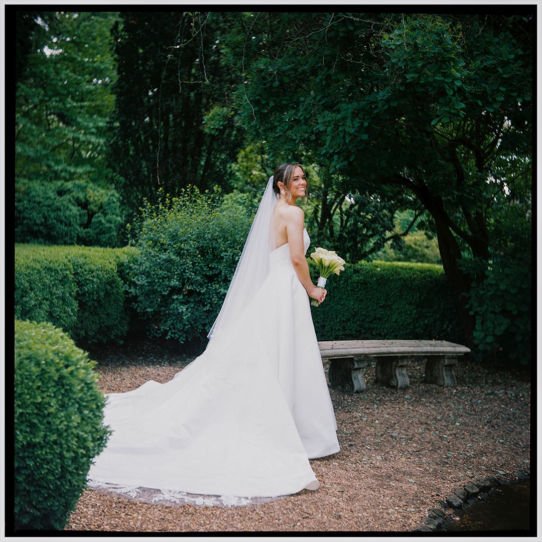 A bride in a flowing white wedding dress and veil walks gracefully through a serene garden setting.