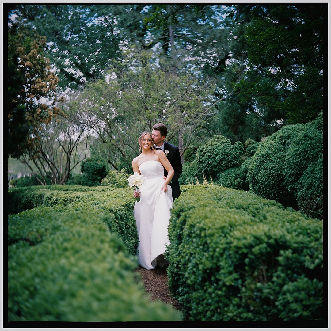 A couple embraces on a garden path surrounded by manicured hedges and lush greenery on their wedding day.