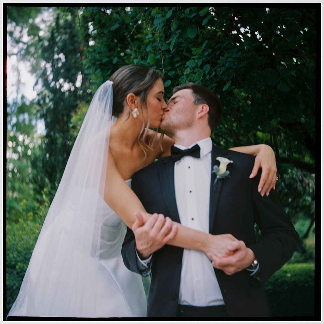 Romantic wedding couple sharing intimate kisses in an outdoor garden setting with lush greenery in the background.